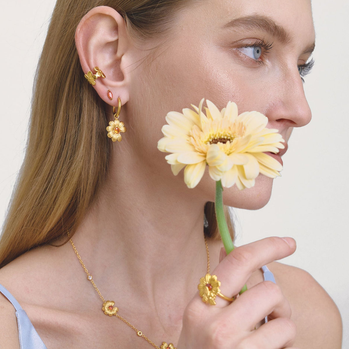 A close-up lifestyle shot of a woman with gold floral jewelry, including a flower-shaped ring, hoop earrings, and a delicate necklace, holding a pale yellow daisy against a clean off-white background.