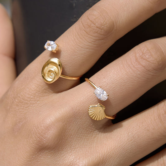 Hand wearing two stacked 18K gold plated stainless steel open cuff rings featuring a spiral nautilus shell, a scallop fan shell, and sparkling oval cubic zirconia stones.