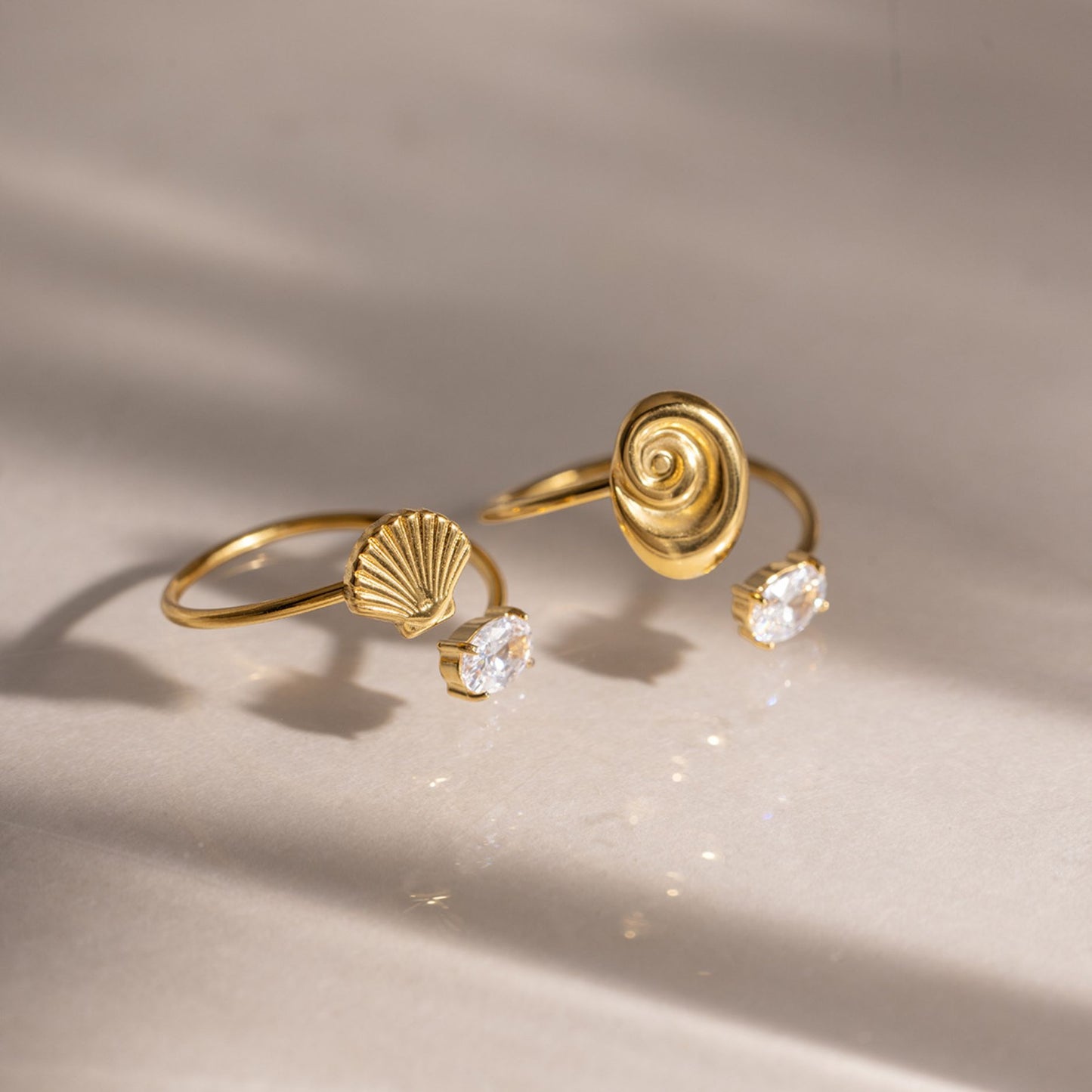 Two waterproof gold shell rings—one spiral nautilus and one scallop fan variant—with cubic zirconia details displayed on a neutral surface under warm summer sunlight.