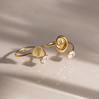 Two waterproof gold shell rings—one spiral nautilus and one scallop fan variant—with cubic zirconia details displayed on a neutral surface under warm summer sunlight.