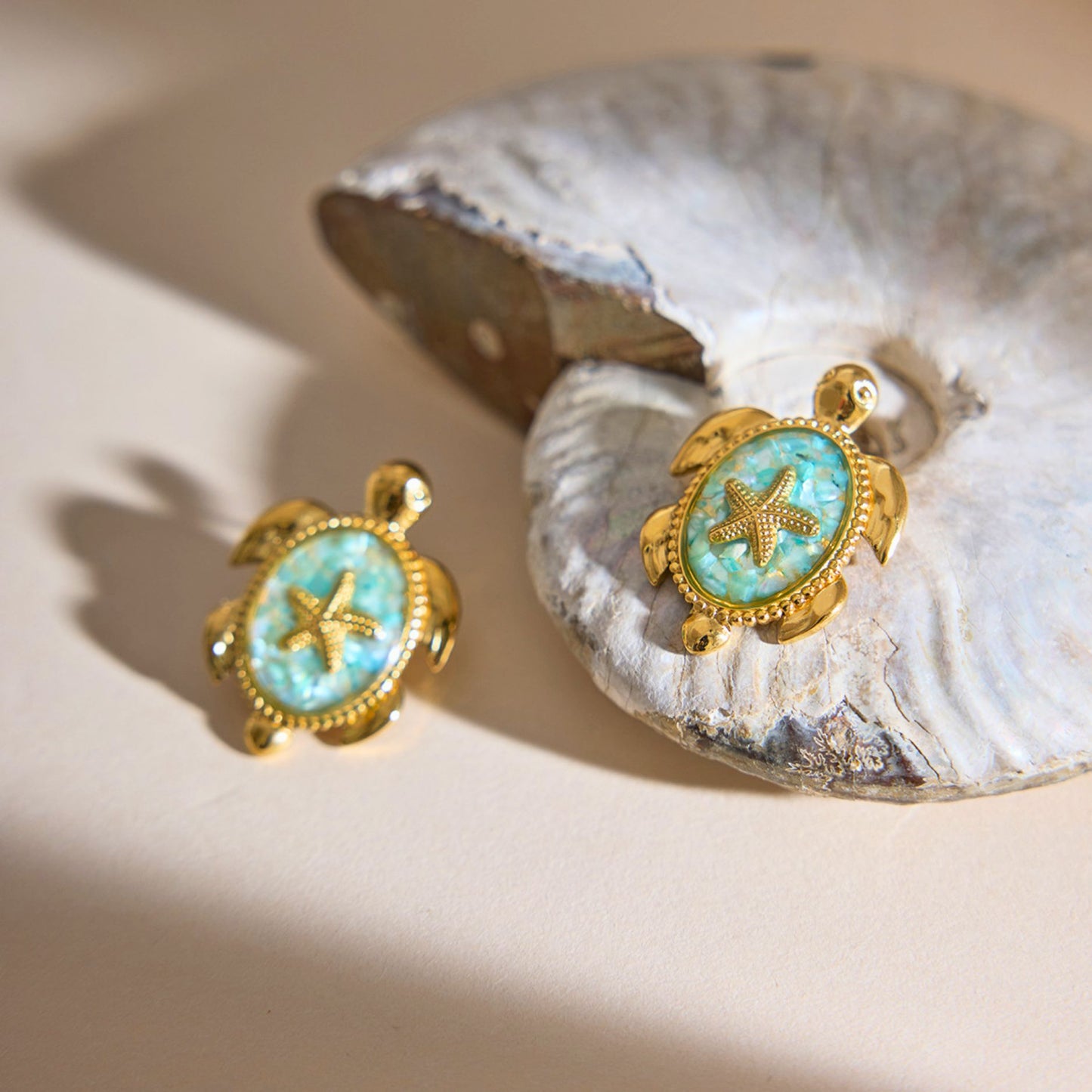 A pair of waterproof gold sea turtle post earrings with bright turquoise textured shells and miniature starfish embellishments displayed on a neutral stone shell background.