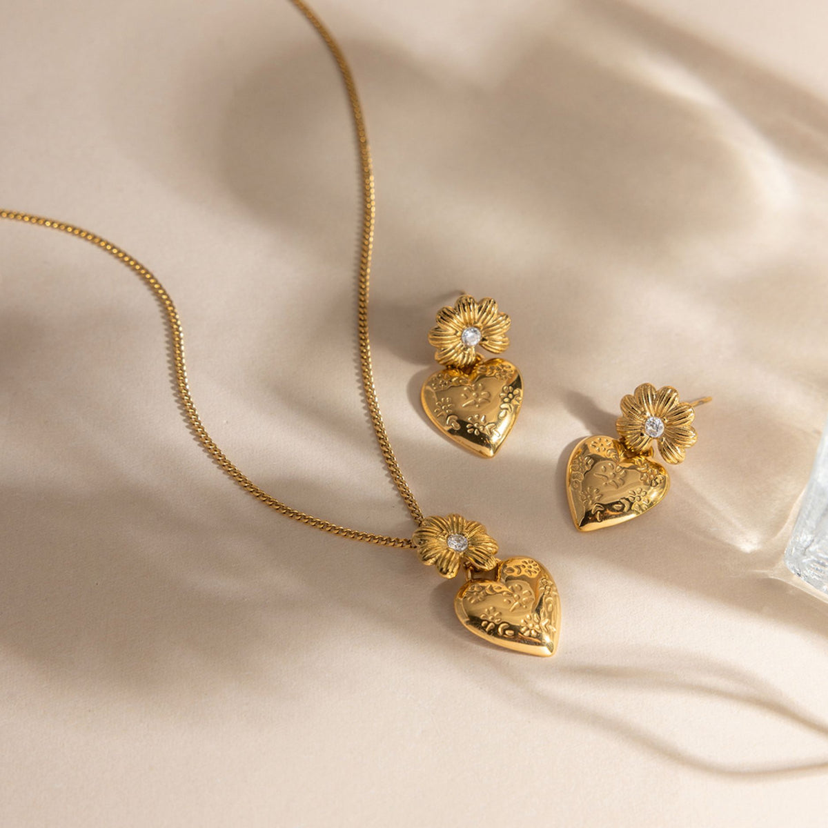 Gold waterproof stainless steel daisy and embossed heart pendant necklace with matching floral drop earrings on warm background