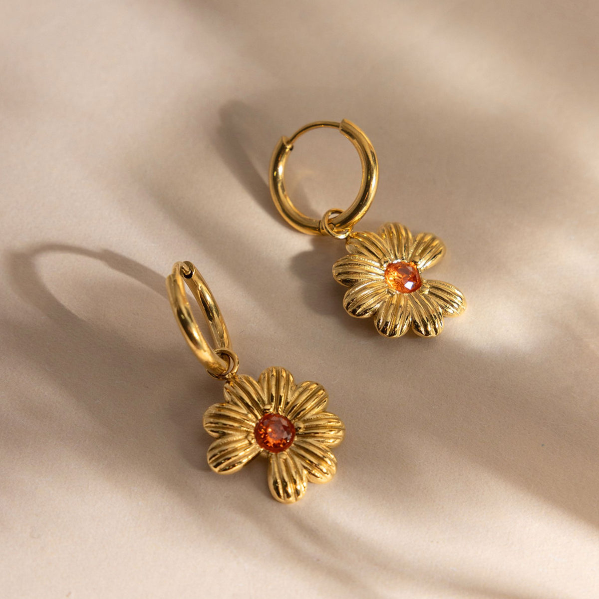 Pair of gold daisy huggie hoop earrings made of tarnish-free stainless steel with warm amber crystal centers on an off-white ceramic dish