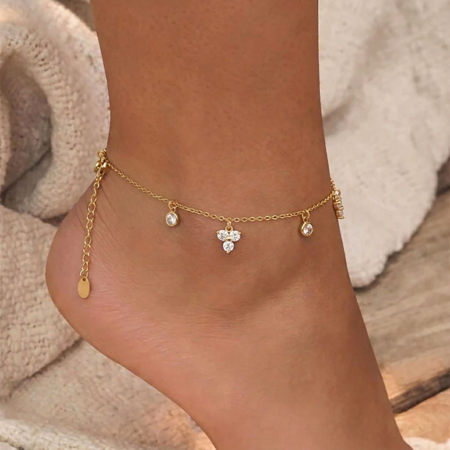 Close up of a model's ankle wearing an 18K gold plated waterproof charm anklet with sparkling CZ crystals against a sandy beach backdrop.