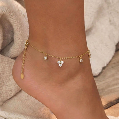 Close up of a model's ankle wearing an 18K gold plated waterproof charm anklet with sparkling CZ crystals against a sandy beach backdrop.