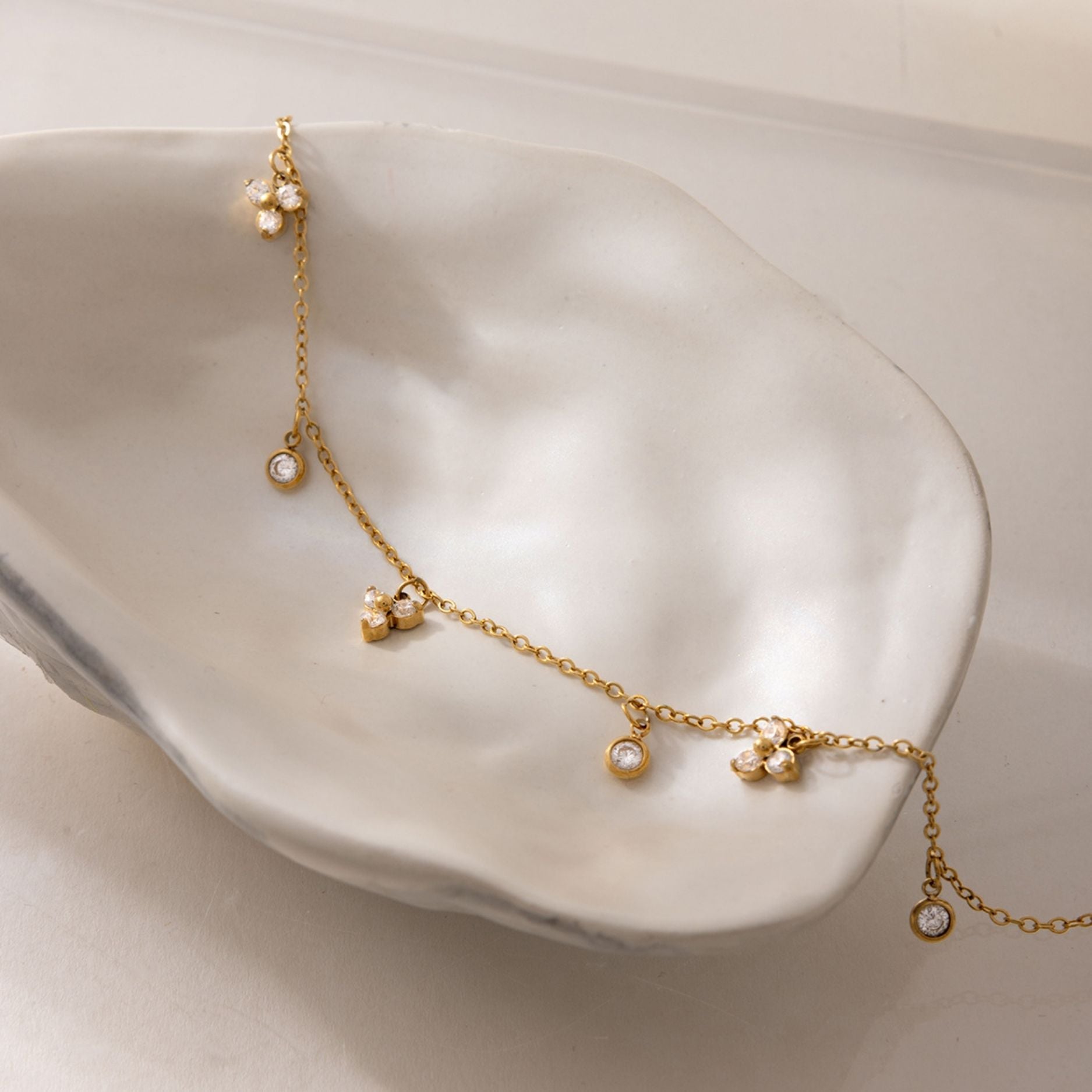A delicate gold stainless steel beach charm anklet with bezel-set crystals resting inside a white porcelain decorative shell