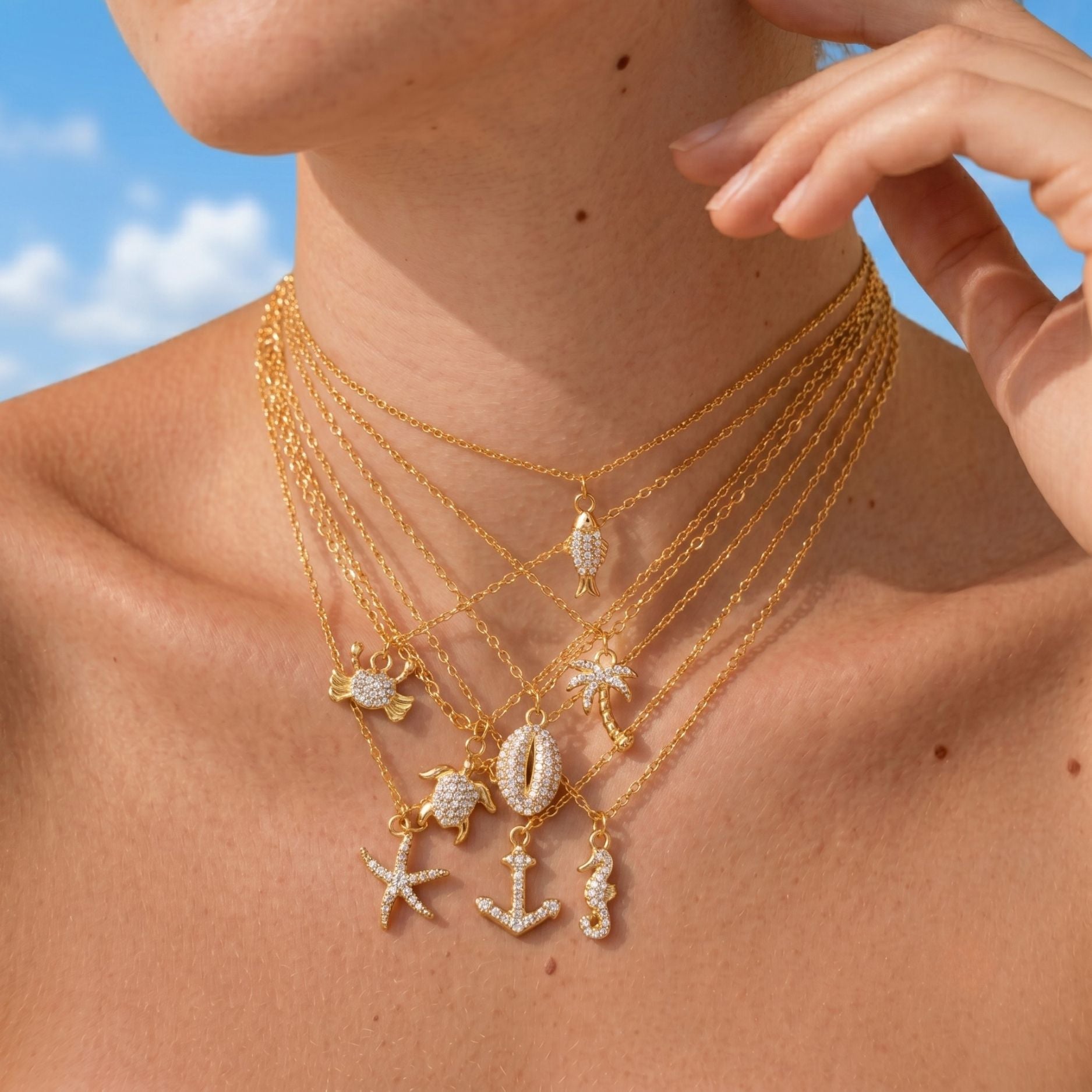 Close-up of a person wearing multiple gold necklaces with various charms against a blue sky. tutle starfish palm